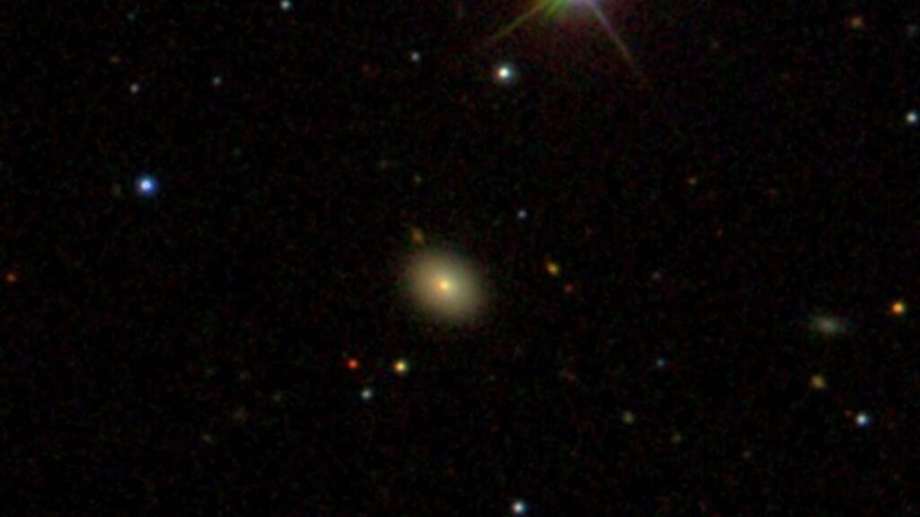 A hazy yellow dot in the center of a dark patch of space. Other colorful dots are around as well.