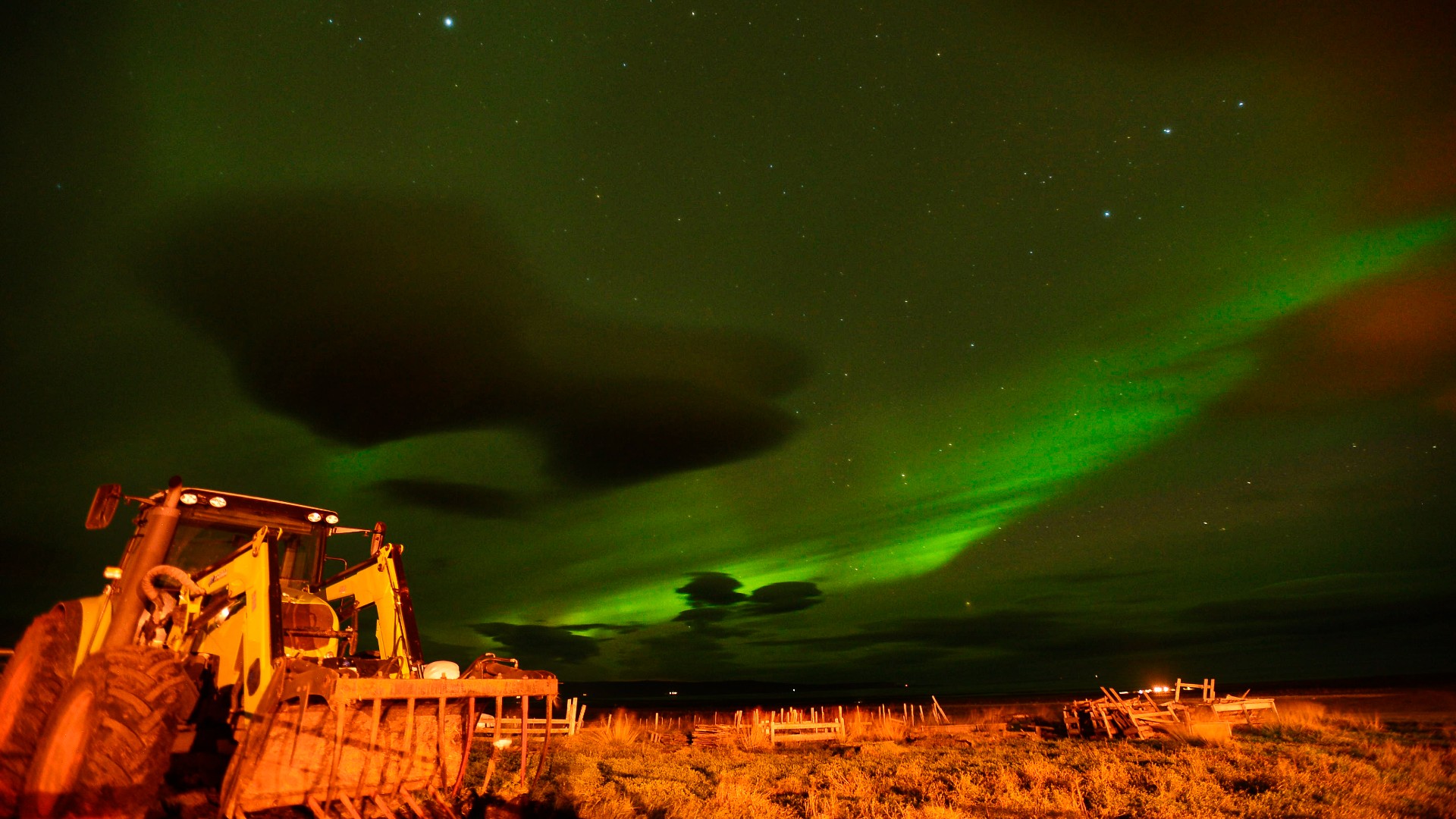 auroras create bands of wispy green light in the sky above a tractor parked in a field on a farm